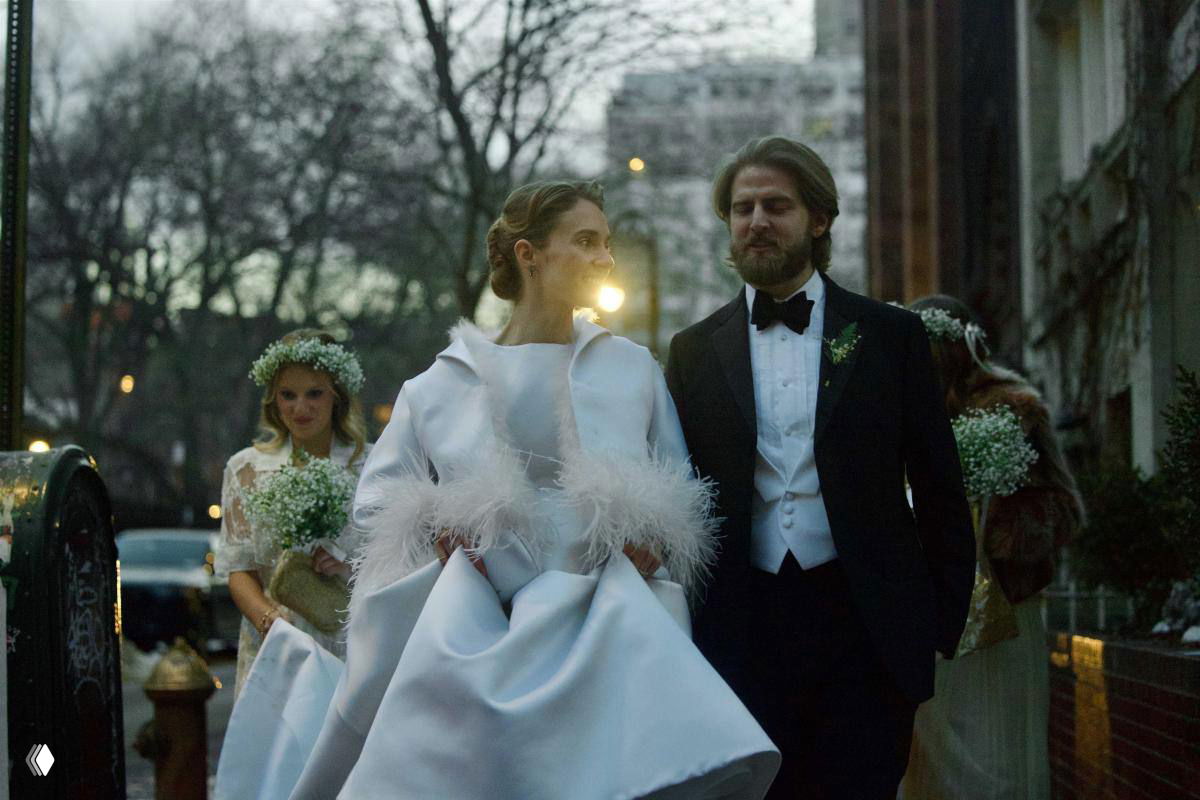 Невеста и жених идут по вечерней улице впереди подружек невесты в венках; на заднем плане городской пейзаж и огни.