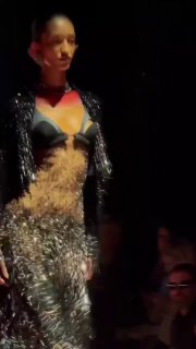 Модель на подиуме в эффектном образе Schiaparelli: блестящее платье с перьями и прозрачными вставками, зрители на заднем плане