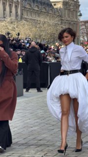 Женщина в полностью белом платье миди с асимметричным подолом и туфлями идет по подиуму у Лувра, вокруг — фотографы и публика.