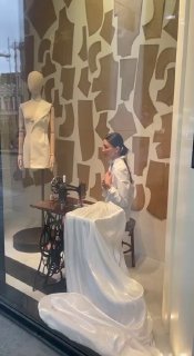 Инсталляция в витрине 12 STOREEZ: сидящая фигура манекена в белом платье с швейной машиной на фоне рулонов ткани и декоративного оформления