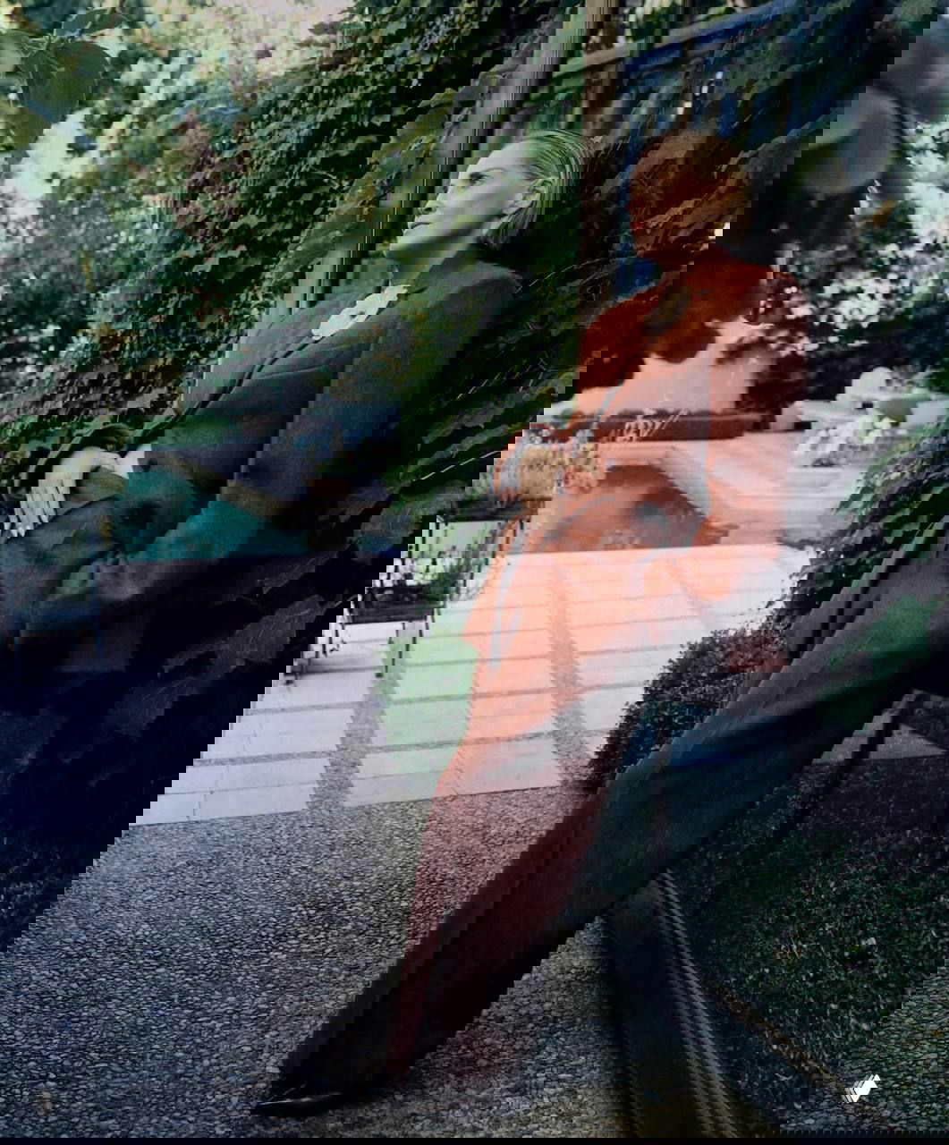 Кадр у бассейна: женщина в кирпично‑коричневом брючном костюме опирается о стену, окружённая садовой растительностью — элегантный образ.