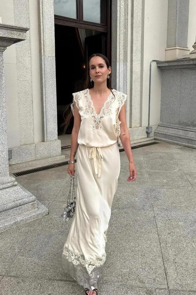 Женщина в кремовом длинном платье с кружевными деталями идёт по парадной зоне; спокойный пастельный образ с нейтральным макияжем.