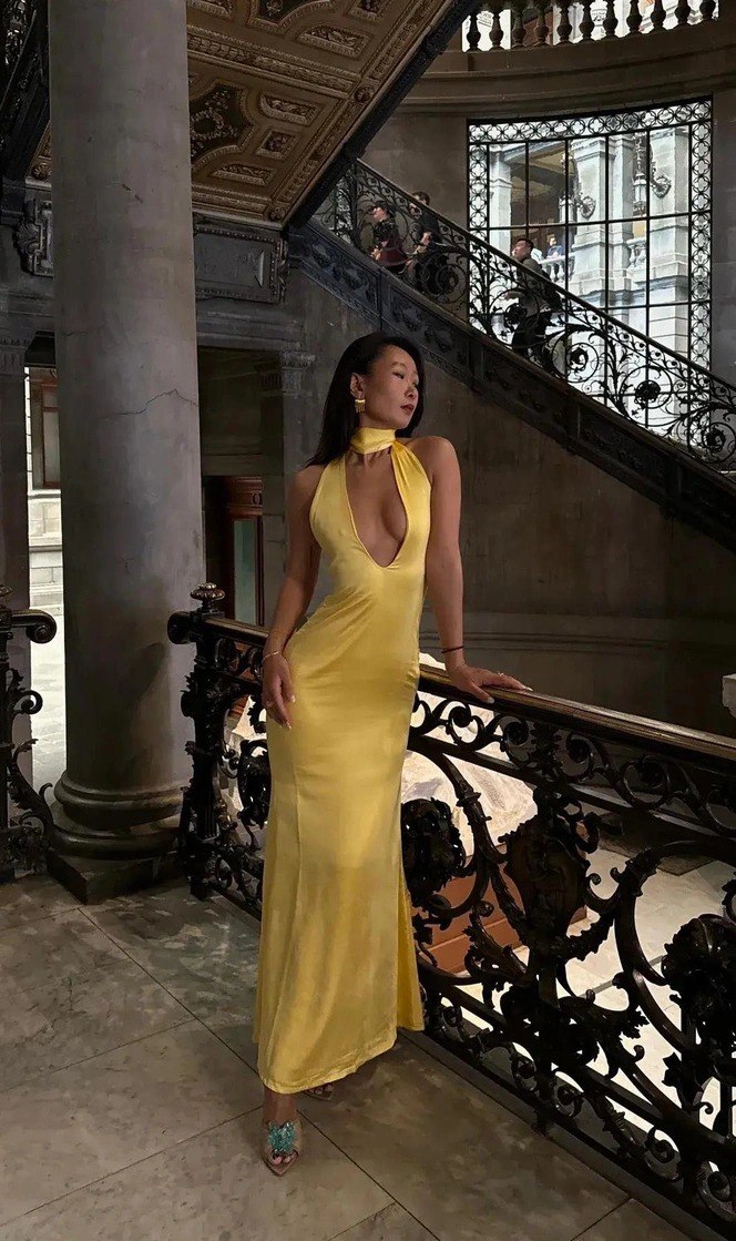 Женщина в длинном жёлтом атласном платье стоит на парадной лестнице старинного интерьера; образ с элегантной обувью и минимальными украшениями.