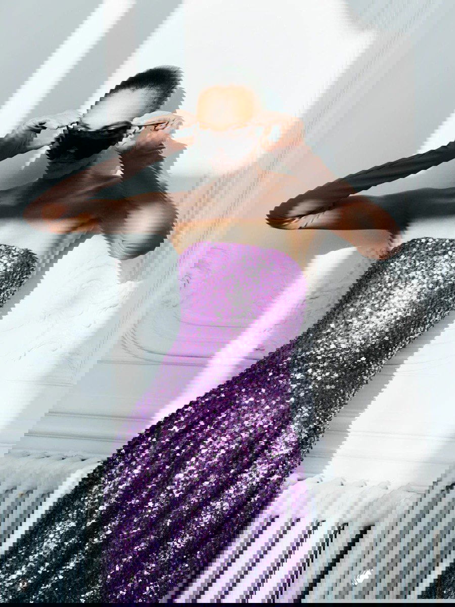 Модель в фиолетовом блестящем платье и солнцезащитных очках стоит в интерьере с лепниной — элегантный студийный кадр коллекции.