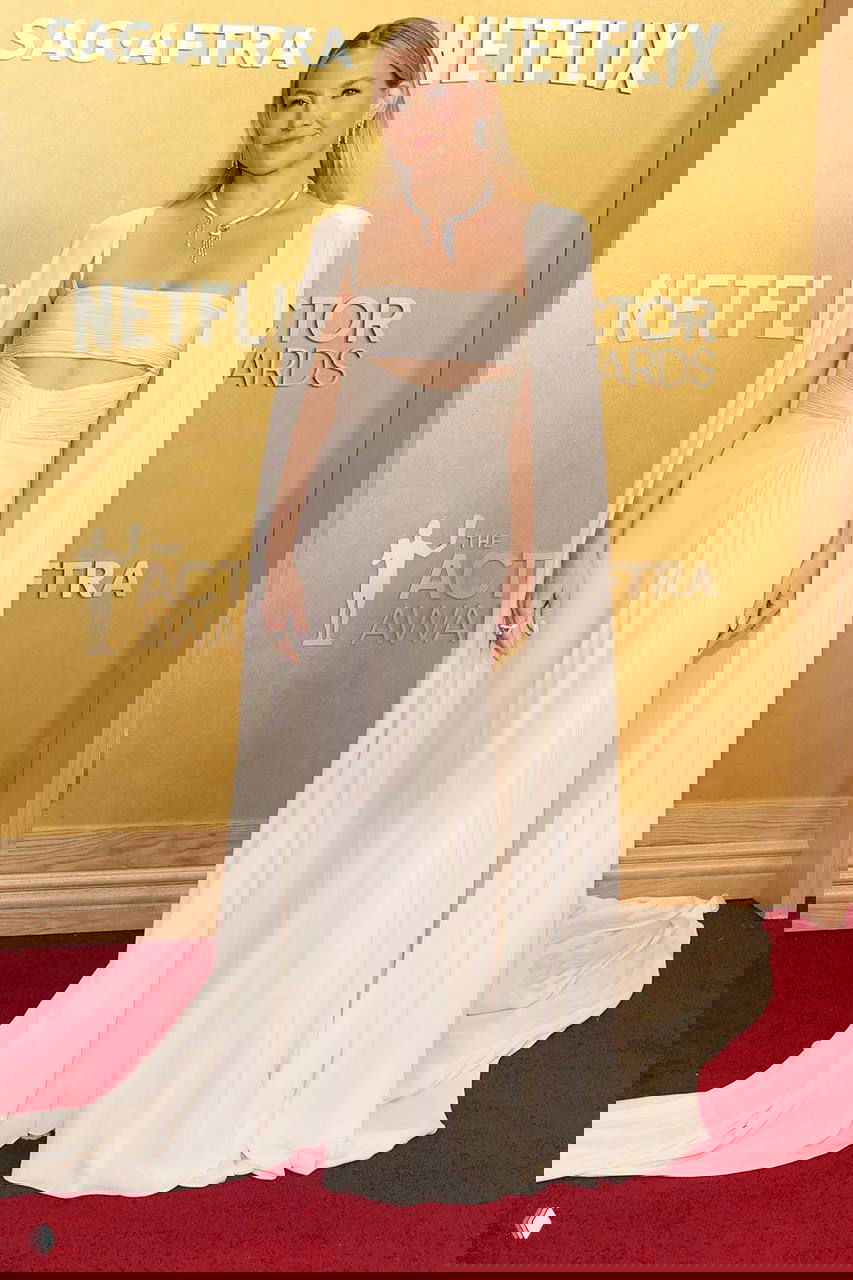 Женщина в кремово-белом струящемся платье с плащевыми элементами на фоне золотой стены и логотипа Netflix/SAG.