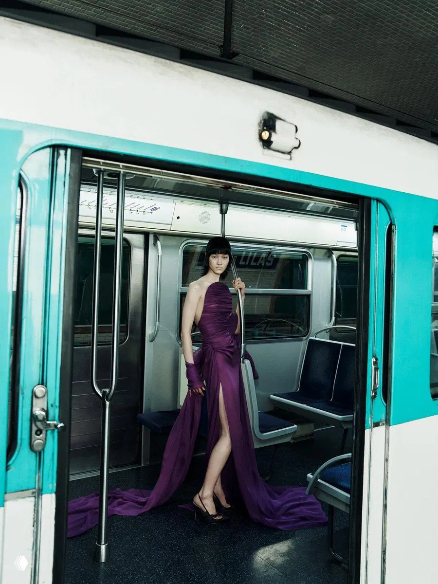 Модель в длинном фиолетовом платье с драпировкой стоит в вагоне метро у поручня — неожиданное сочетание высокой моды и городского пейзажа.