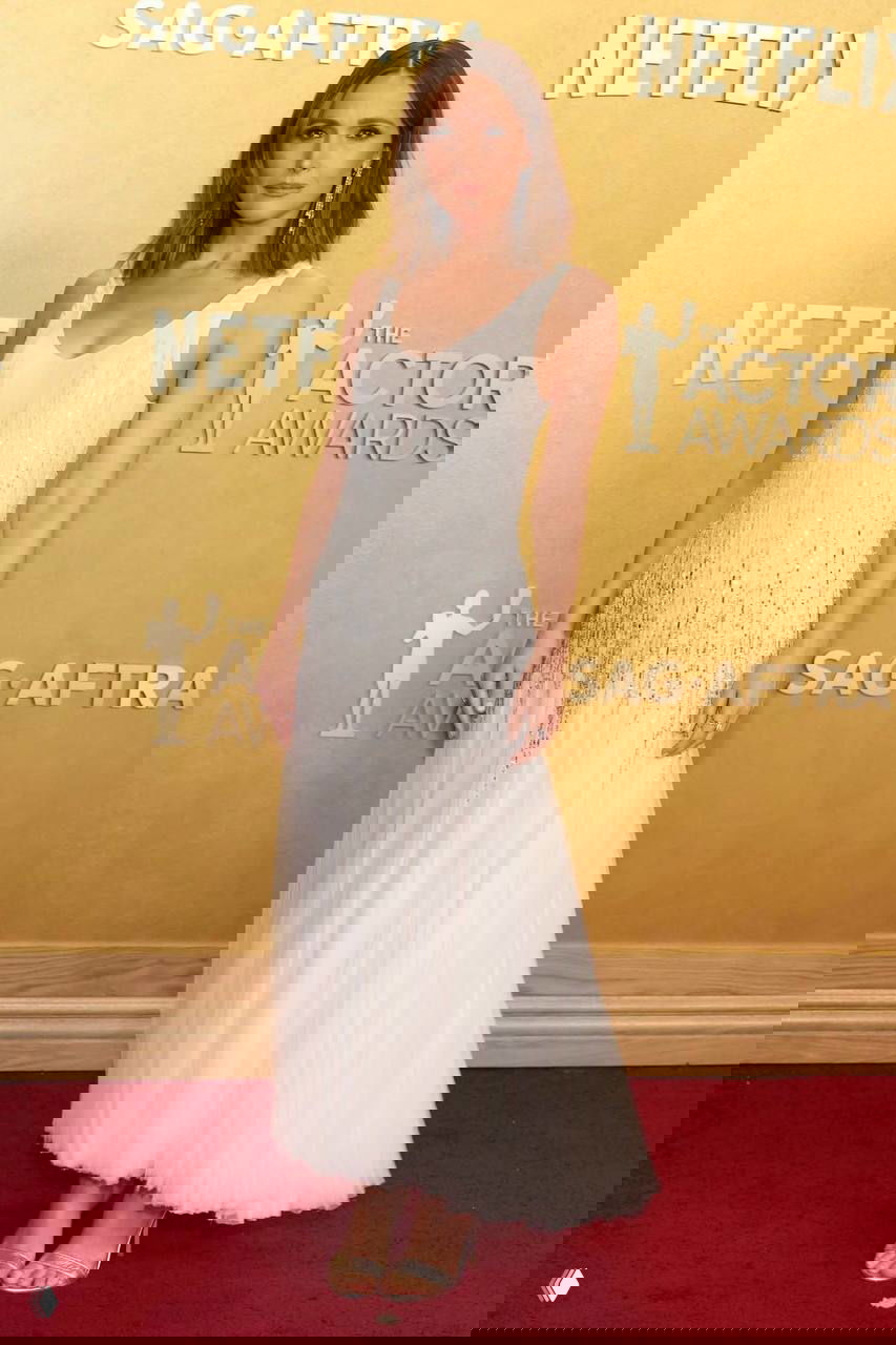 Женщина в лёгком белом платье с бликами и плиссировкой на фоне золотой бренд-стены премии, видно обувь и красный ковер.