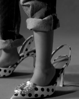 Крупный план чёрно‑белого снимка: детские ноги в больших туфлях Amina Muaddi с гороховым принтом и декоративной пряжкой.