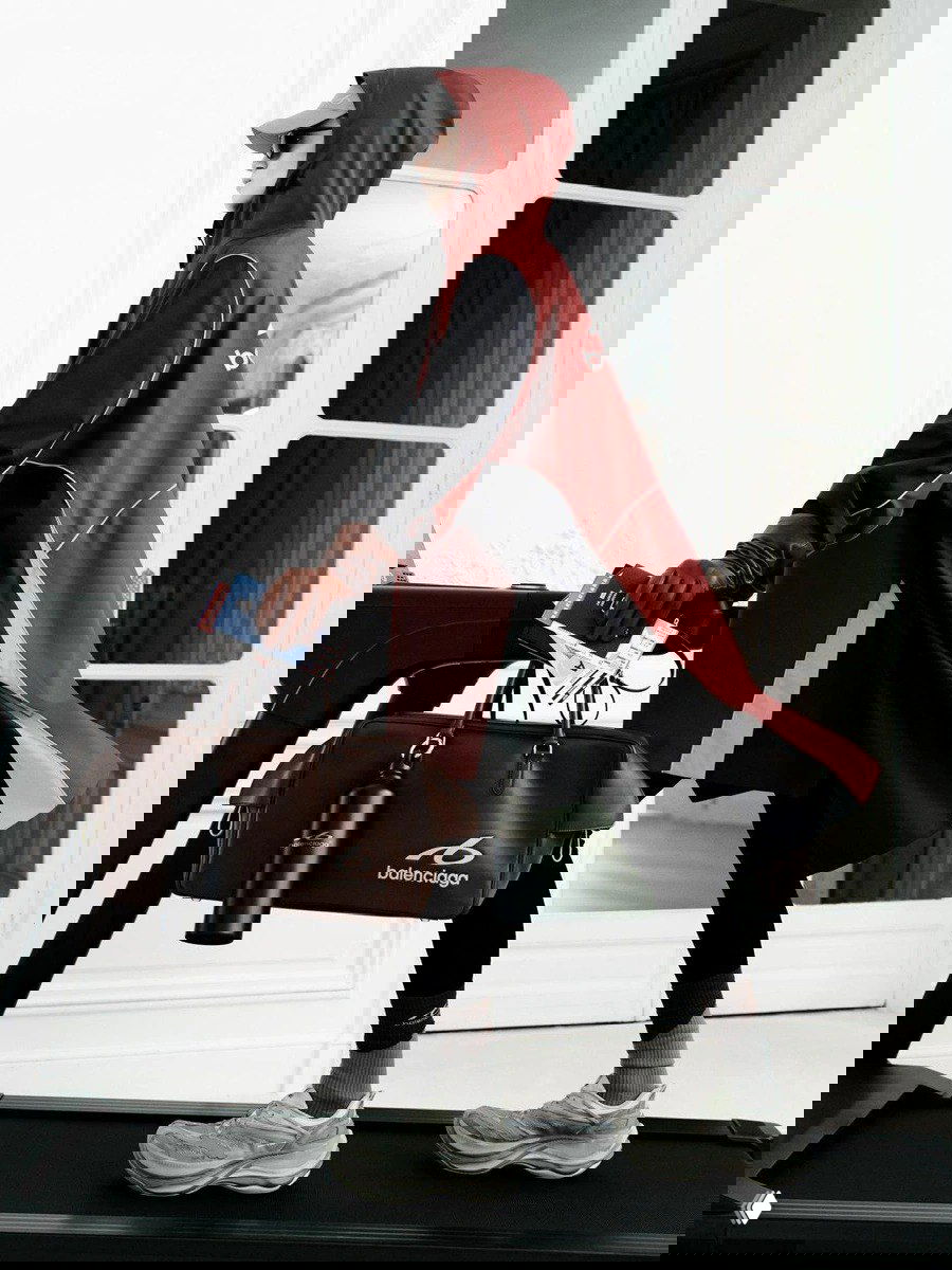 Модель в красно-чёрной парке с капюшоном и спортивной сумкой Balenciaga идёт по коридору, демонстрируя утилитарный спортивный стиль бренда.