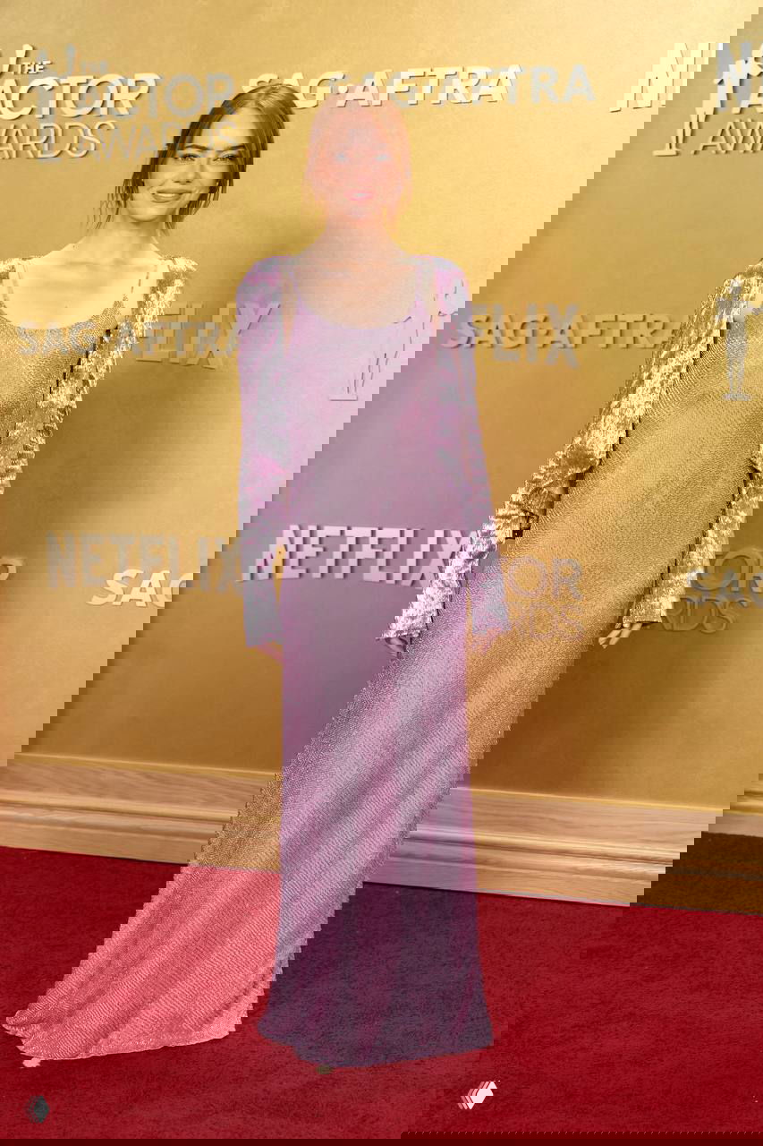 Молодая женщина в фиолетово-сиреневом длинном платье спокойного кроя позирует на красной дорожке премии на фоне золотой фотозоны.