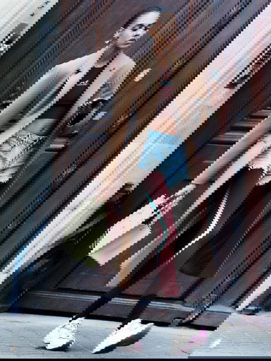 Модель в бежевом пальто, укороченных джинсовых шортах и бордовых леггинсах идёт по городу, сочетая повседневность и люксовый стиль в образе бренда.