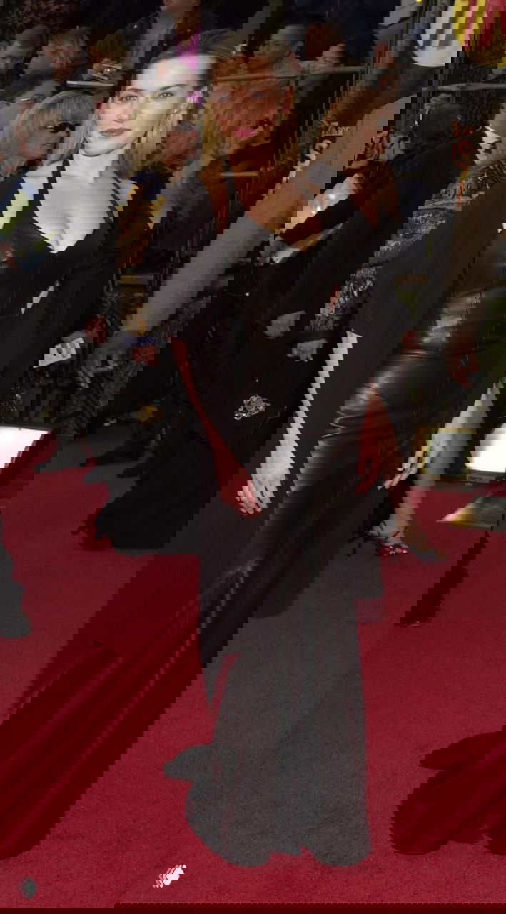 Приталенное кожаное чёрное платье в пол со свободной накидкой на плечах на фоне красной дорожки и гостей мероприятия.