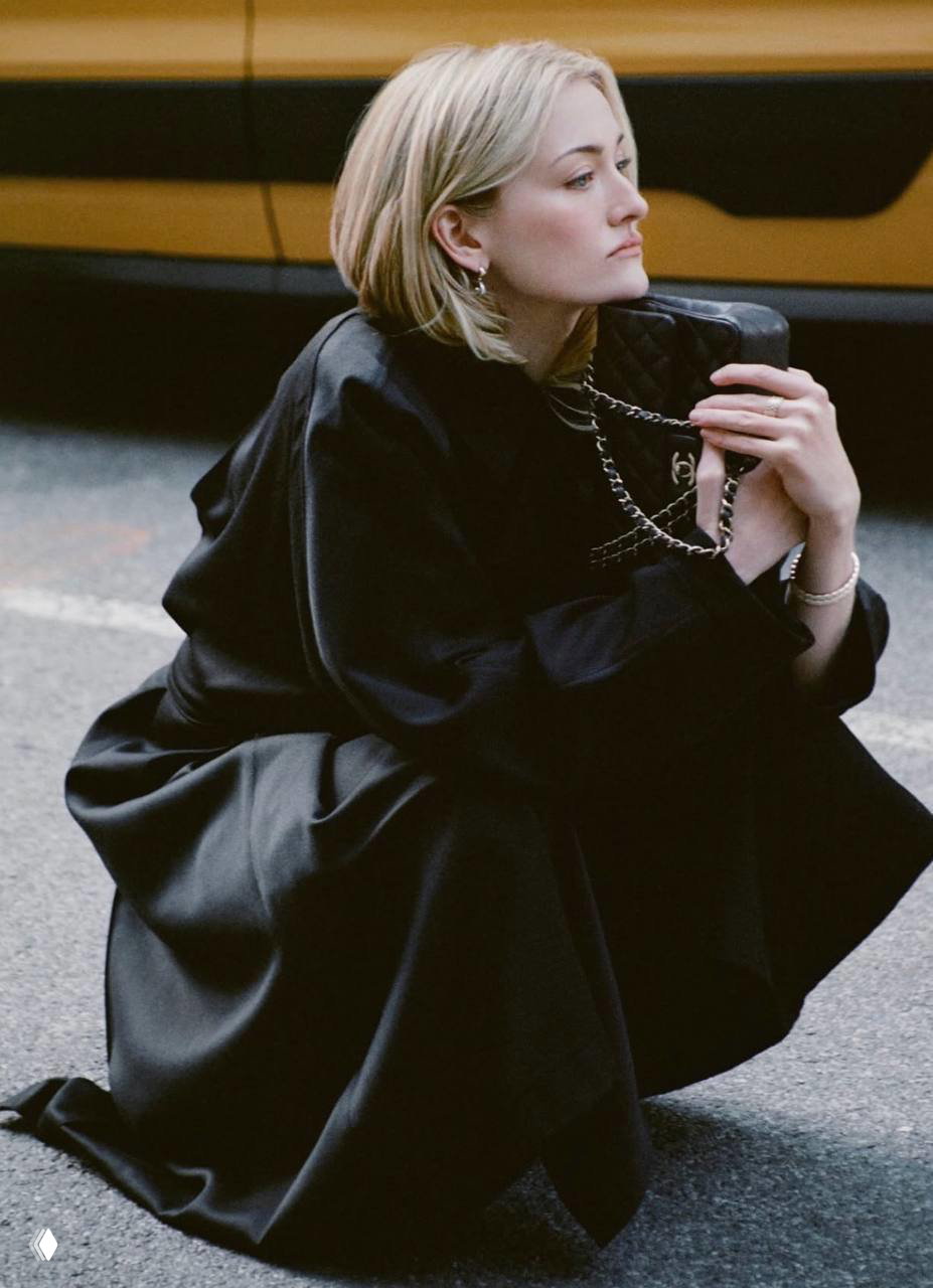 Сидящая Сара Пиджон в чёрном наряде прижимает сумку Chanel к груди, уличный кадр с такси и городской текстурой в фоне.
