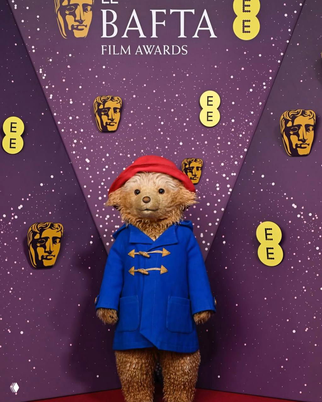 Мишка Паддингтон в полный рост в синем дафлкоте и красной шляпе перед брендированной стеной BAFTA на красной дорожке