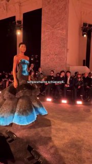 Платье Schiaparelli с объёмными слоями и синими акцентами на подиуме; сцена шоу с подсветкой и внимательной публикой в зале.