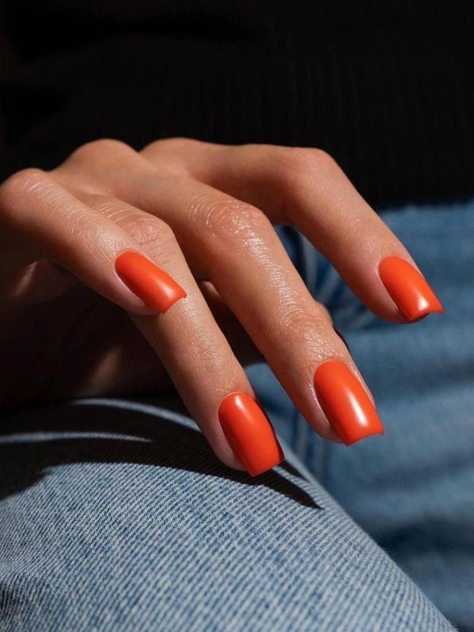 Матовый ярко-оранжевый маникюр на квадратных ногтях, рука расположена на джинсовой ткани, модный смелый оттенок для праздника.