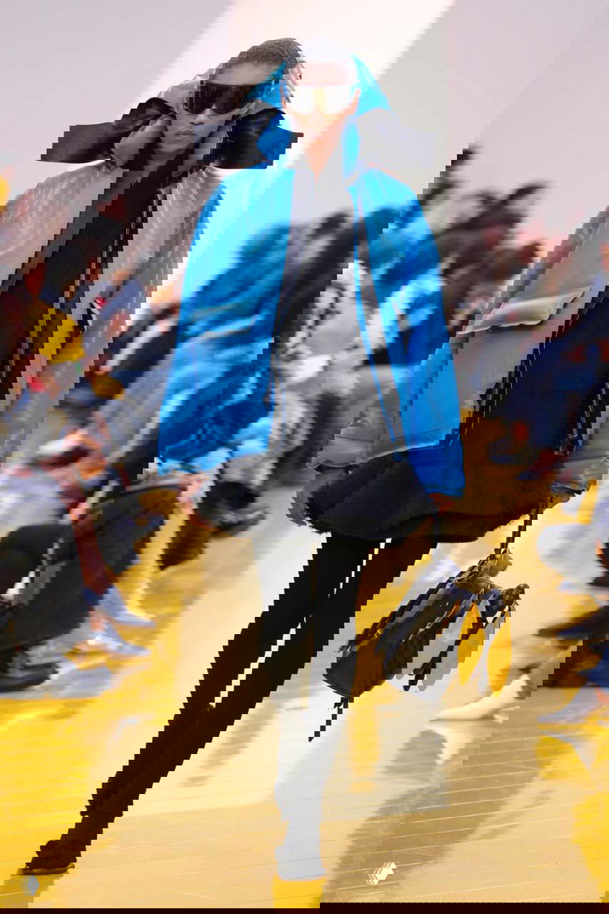 Модель в голубой объёмной куртке с капюшоном и крупными декоративными бантами, тёмные очки и контрастные аксессуары на подиуме Loewe.