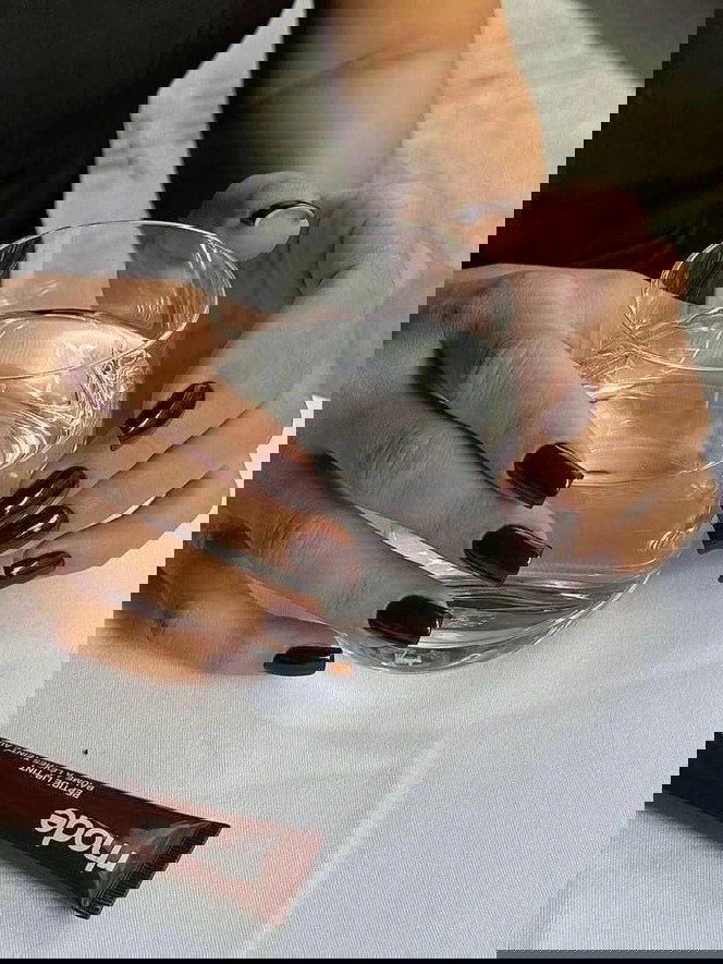 Руки с глубоким шоколадным маникюром держат прозрачный стакан воды, фокус на блеске лака и форме ногтей в дневном свете.