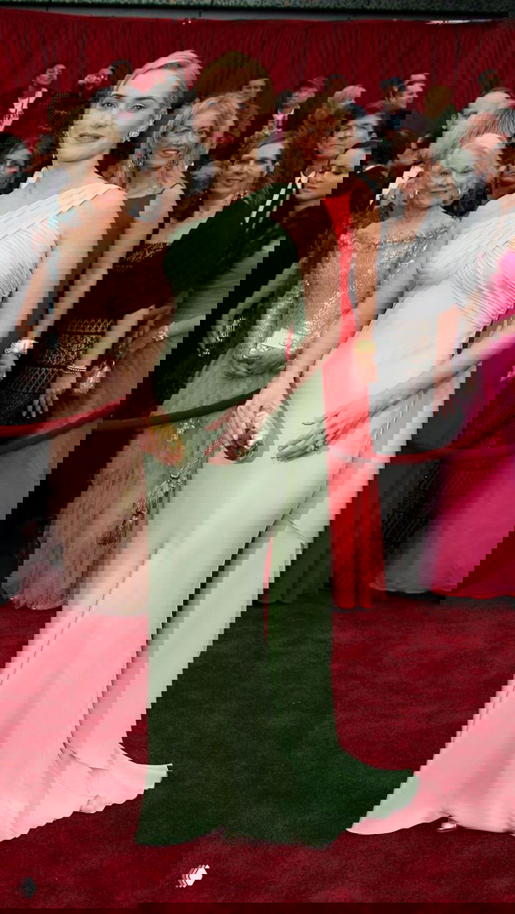 Платье нежно-мятного оттенка на одно плечо с плавными драпировками, стоящая фигура на красной дорожке и зрители позади.