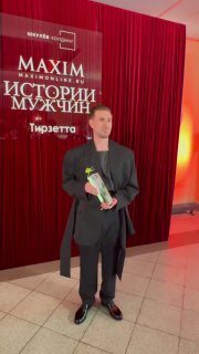 Мужчина в чёрном костюме держит цветы на фоне красной занавески и баннера MAXIM «Истории мужчин» в фойе Исторического музея.