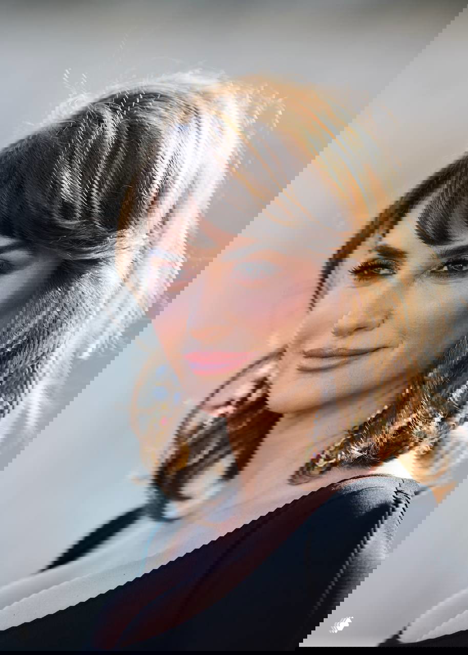 Крупный план женщины с выраженным макияжем и серьгами, видны верх платья и глубокое декольте на пресс-мероприятии.