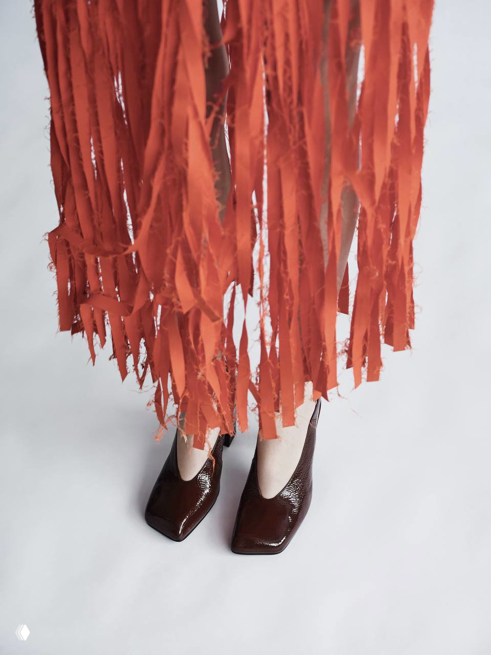 Кадр снизу: ноги модели в лаковых коричневых туфлях с квадратным носком на фоне оранжево‑рваной бахромчатой юбки, минималистичный студийный фон.