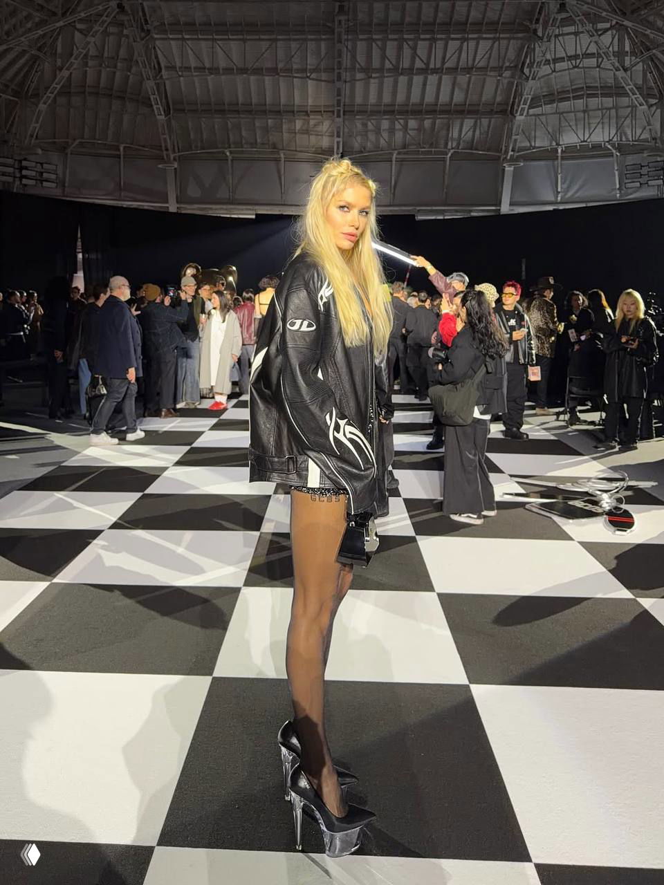 Лена Перминова в кожаной куртке на фоне шахматного подиума и зрителей — атмосферный кадр с показа и толпа вокруг.