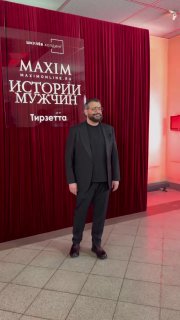 Бородатый мужчина в тёмной одежде на фоне красной занавески и баннера MAXIM «Истории мужчин» в фойе музея — кадр с гостем мероприятия.