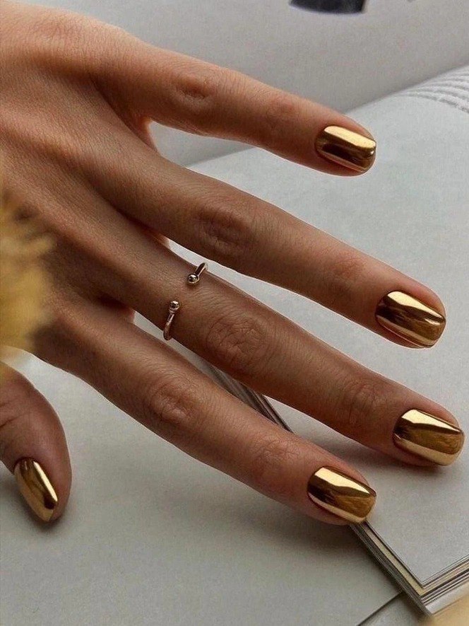 Крупный план руки с зеркальным золотым хром-маникюром на коротких ногтях, отражения и металлический блеск создают роскошный праздничный акцент.