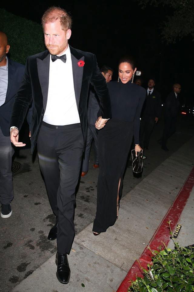 Пара идёт по тротуару вечером: мужчина в смокинге и женщина в длинном чёрном платье, фото сделано папарацци при выходе с мероприятия.