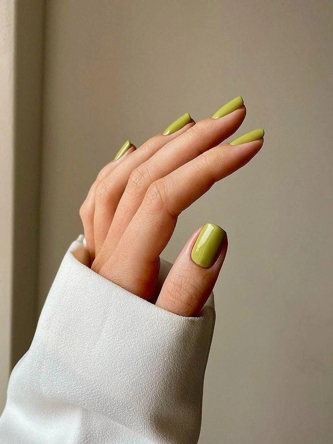Рука с салатово-зелёным маникюром на аккуратных квадратных кончиках ногтей, светлый нейтральный фон подчёркивает свежесть оттенка.