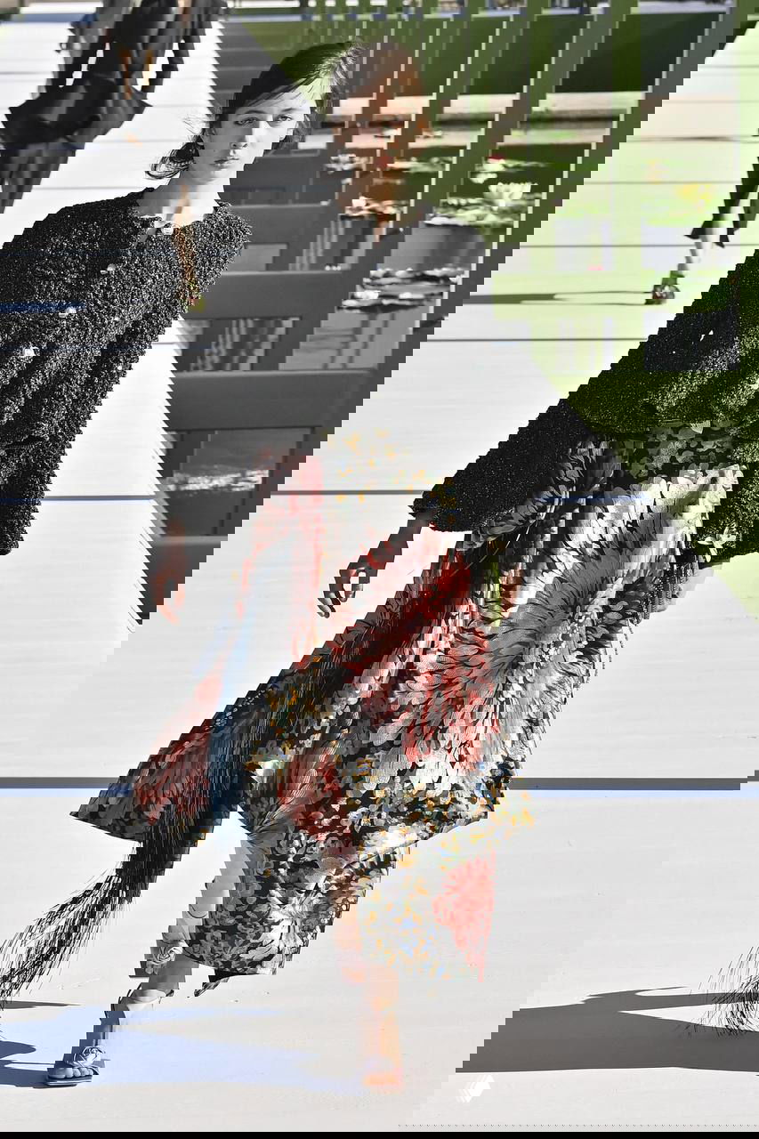 Модель в черной вязаной куртке и широкоматовой юбке с крупным цветочным принтом; кадр подчёркивает декоративный рисунок и силуэт одежды на подиуме.