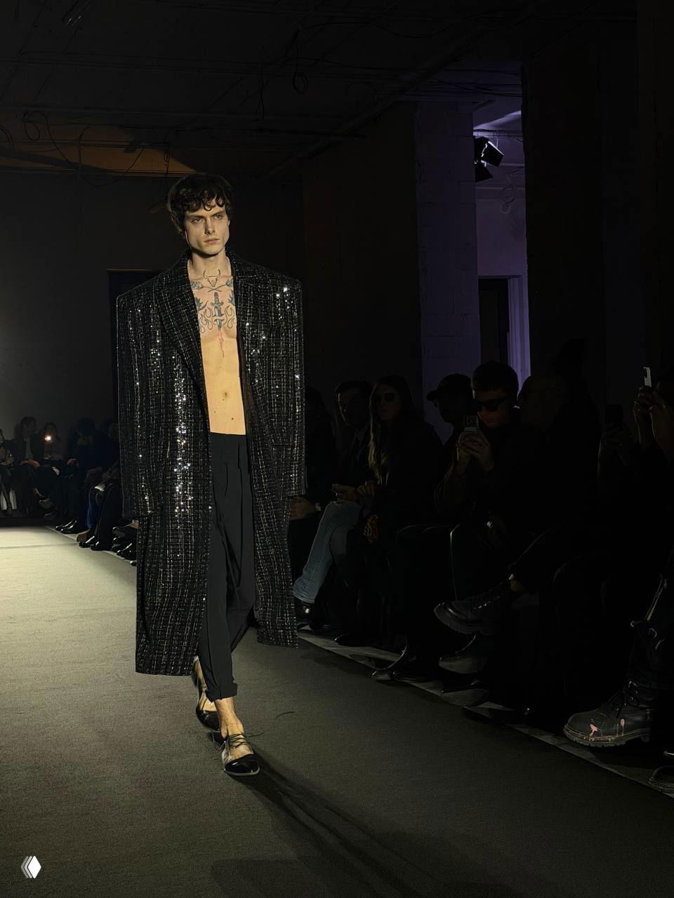 Мужская модель в длинном блестящем пальто с оголенным торсом и татуировками, контрастный рок-образ на подиуме Viva Vox.