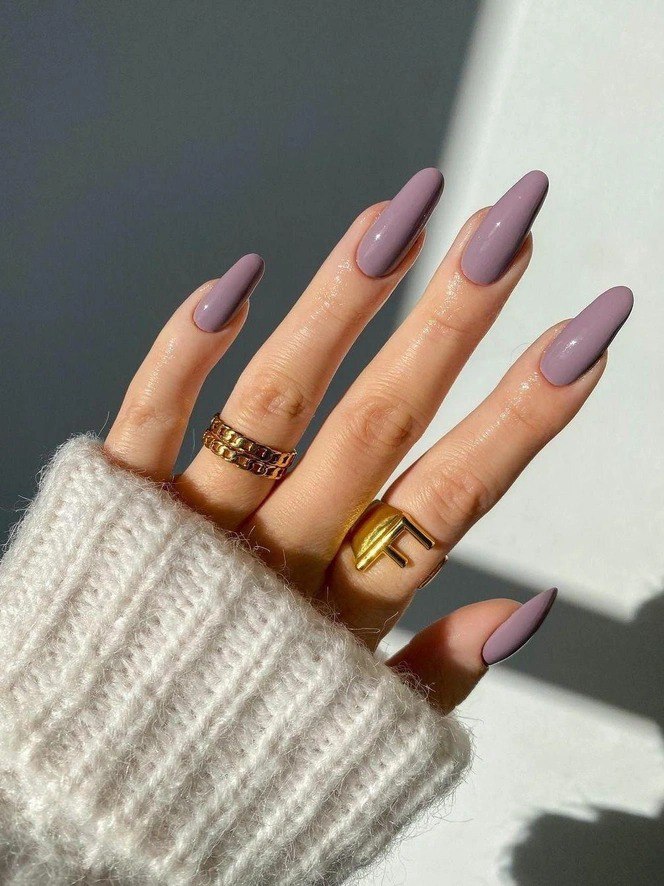 Рука с пастельно-лиловым маникюром и золотыми кольцами на пальцах, мягкий свет и вязаный рукав создают уютный стильный образ.