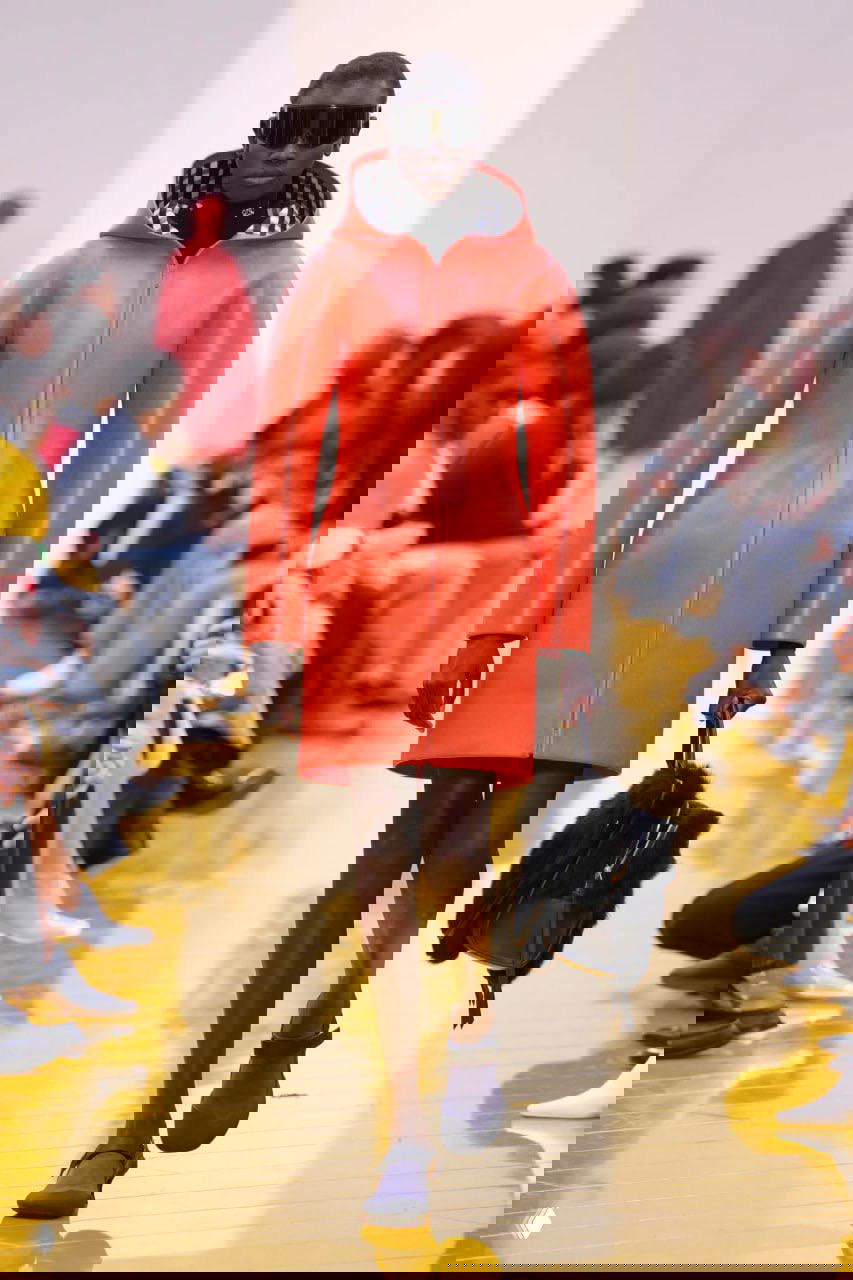 Модель в красном гладком пальто с капюшоном, обтекаемые линии и минималистичная форма сочетаются с крупной сумкой, акцент на силуэт.