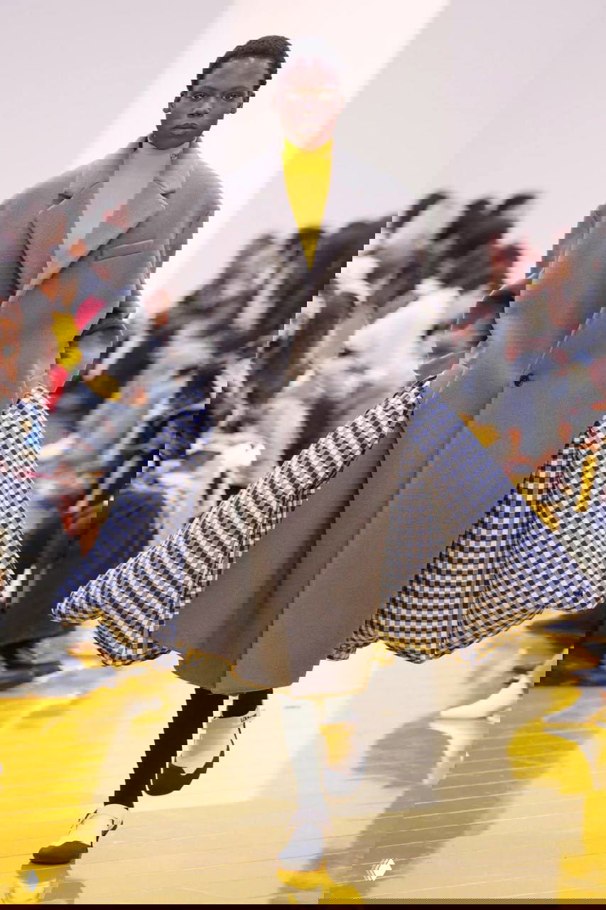 Оверсайз‑пальто серого цвета с открывающимся клетчатым подплечьем, внутри — клетчатая подкладка; образ завершён жёлтым свитером и сдержанными аксессуарами.