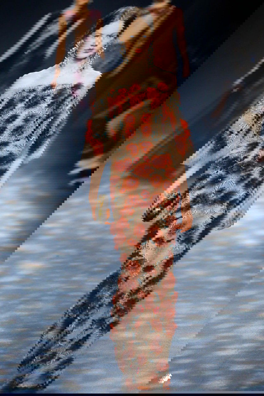 Длинное платье Marchesa с прозрачной основой и обильной текстурной отделкой в тёплых красно‑коричневых тонах на подиуме.