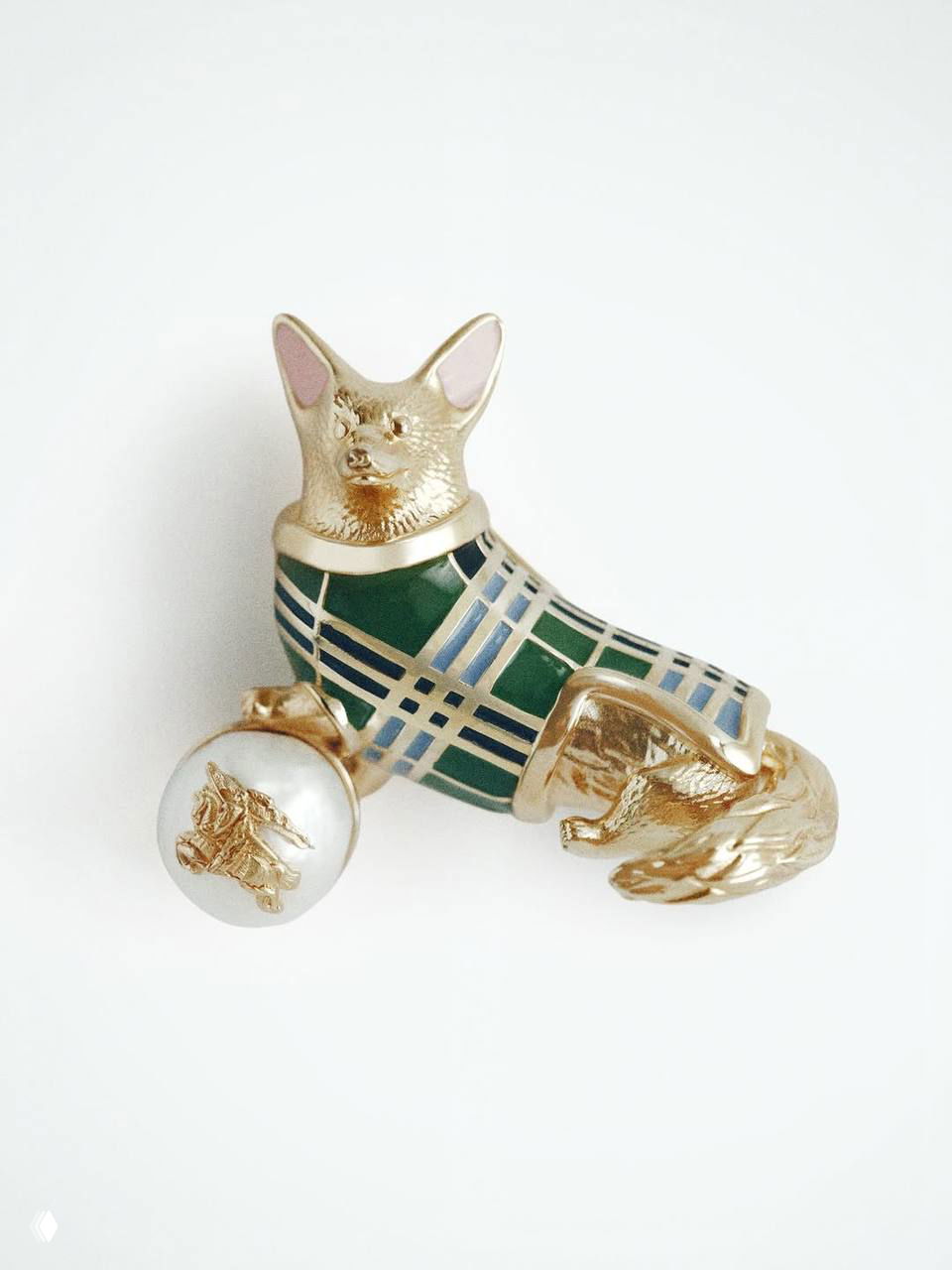 Крупный план броши‑корги от Burberry: золотистая эмалевая фигурка корги в тартановом наряде, декоративный шар рядом.