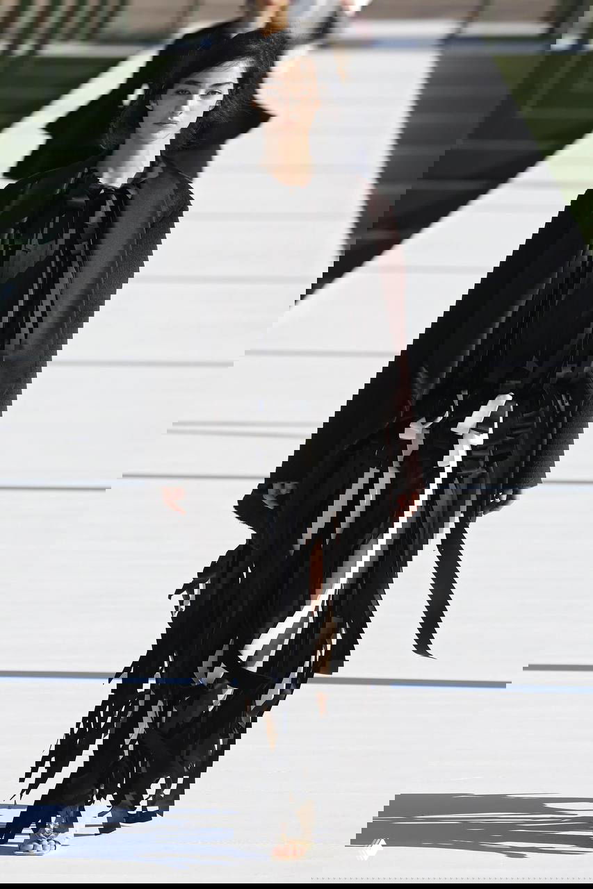 Чёрное платье с длинными лентами и широкий пиджак: драматический образ с акцентом на движение ткани и силуэт, показанный на светлом подиуме на фоне декоративных рам.