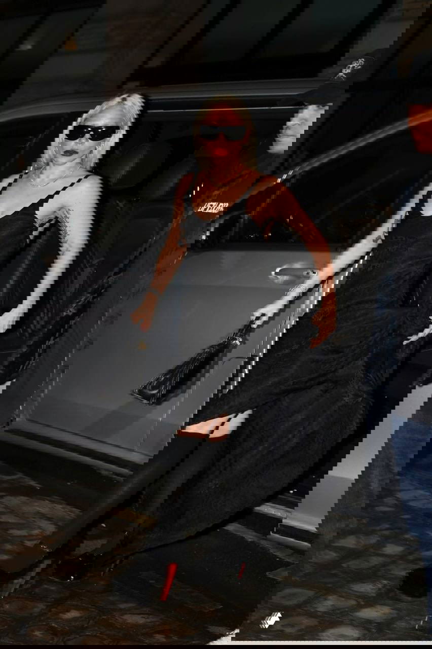 Ночной кадр у автомобиля: чёрное вечернее платье с минималистичным кроем и высокие платформы на влажном тротуаре.