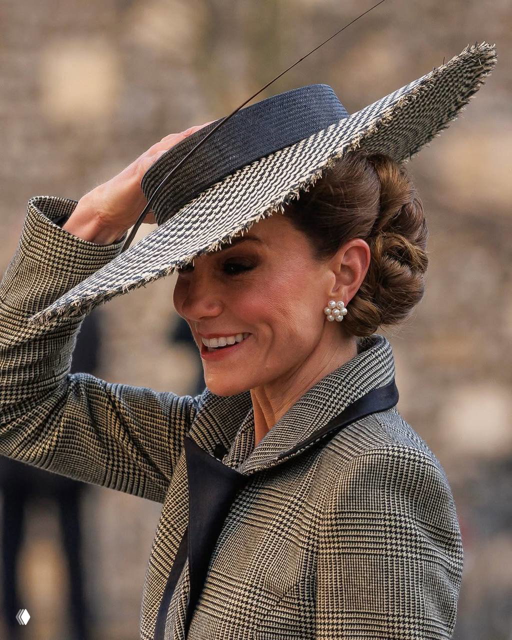 Женщина в твидовом пальто держит широкополую соломенную шляпу рукой и улыбается, уличный портрет с размытым фоном и естественным светом.