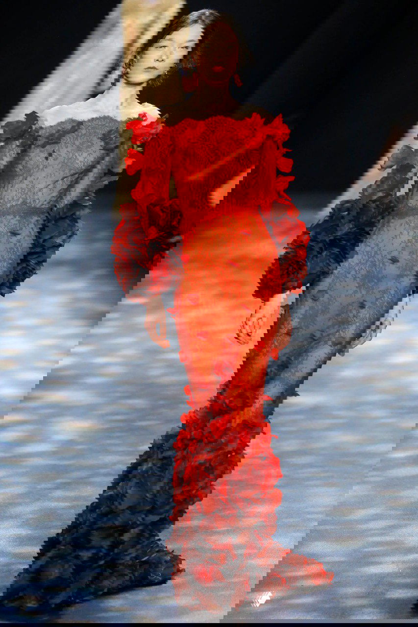 Тонкое алое платье Marchesa с кружевной фактурой и объёмными лепестковыми деталями по подолу и рукавам на подиуме.