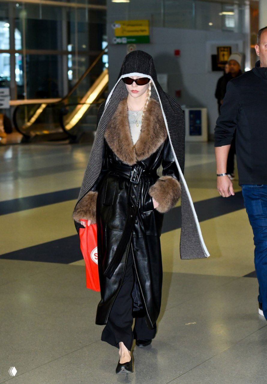 Кадр в ресторане или на выходе: длинное тёмное пальто, повседневные ботинки и сумка через плечо на фоне уличных растений и людей.