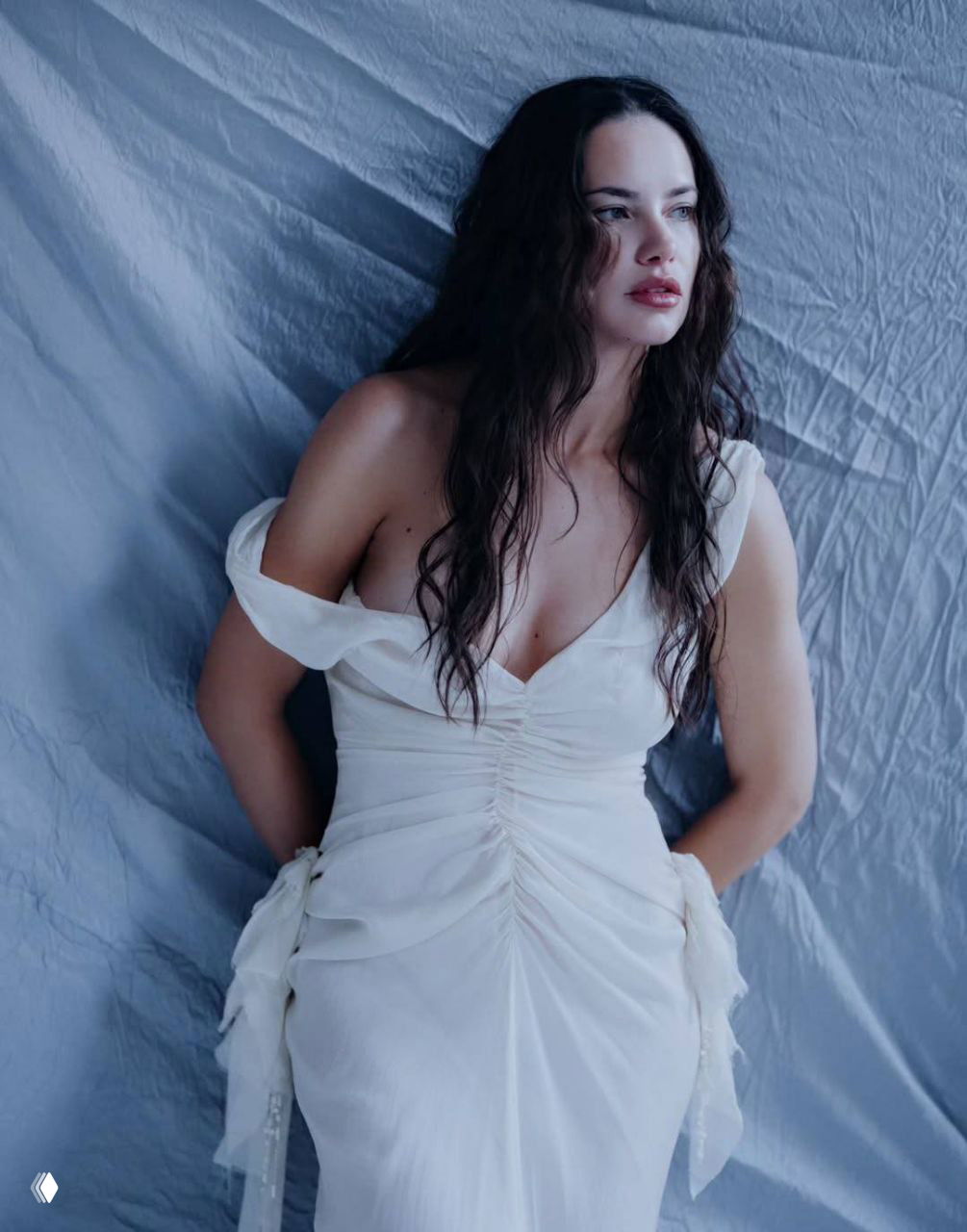 Полноформатный портрет модели в струящемся светлом платье на фоне ткани спокойного голубого оттенка; влажная укладка и естественный макияж.