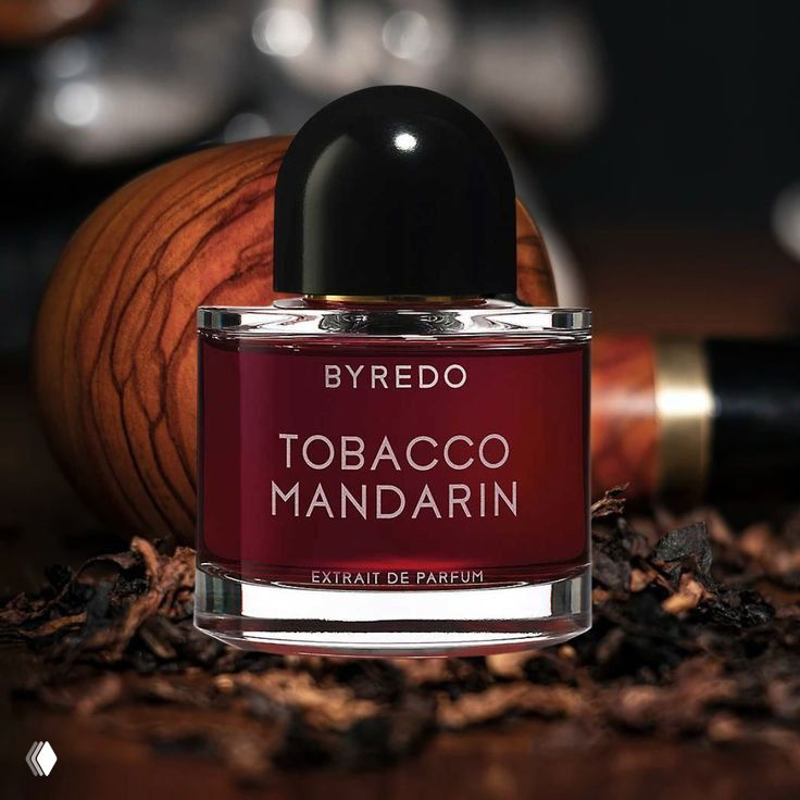 Флакон Byredo Tobacco Mandarin тёмно‑бордового цвета на фоне тёмной текстуры и кофейных зёрен — подчёркнуто тёплая и пряная подача.