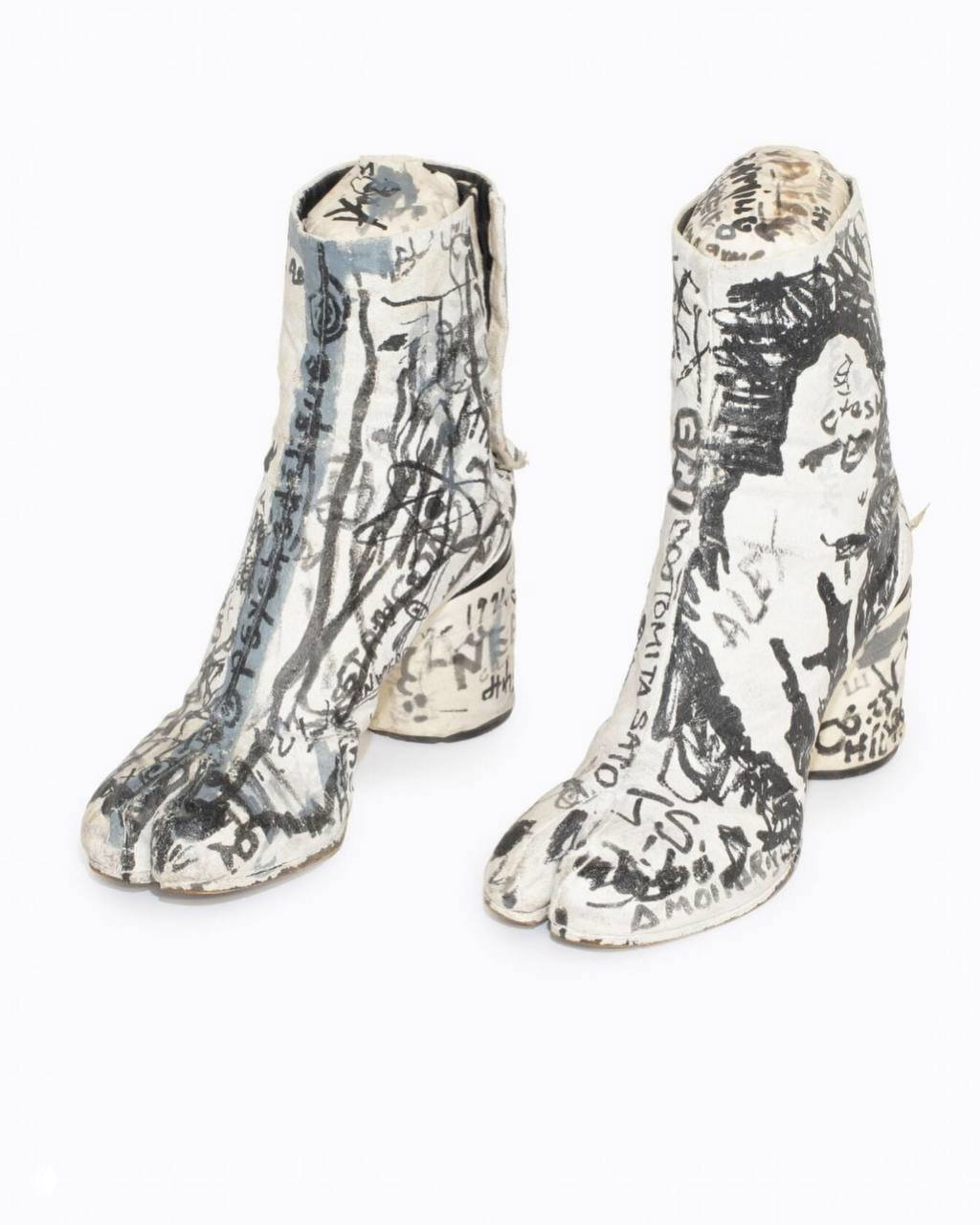 Пара белых таби Maison Margiela, покрытых ручными граффити и надписями, выставленная как лот с белым фоном и видимыми текстовыми каракулями по поверхности