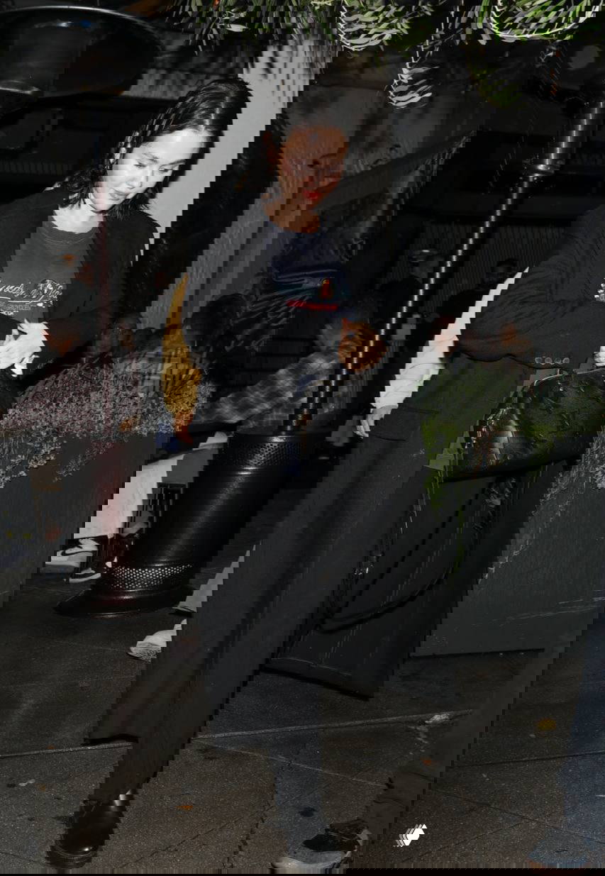 Папарацци‑фото на улице: длинное чёрное пальто, свободный силуэт и сумка‑тоут в руках, спокойный городской фон.