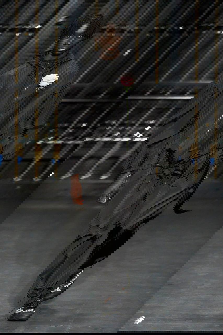 Тёмный нейлоновый костюм с затягивающимися кулисками и приколотой розой — пример «красивой маскулинности» в мужских образах Simon Rocha.