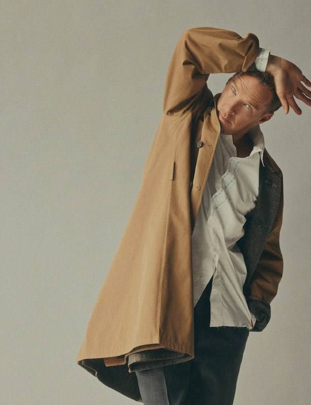 Актёр в длинном плаще в студии: полуназад, художественная поза и мягкое освещение в модной съёмке.