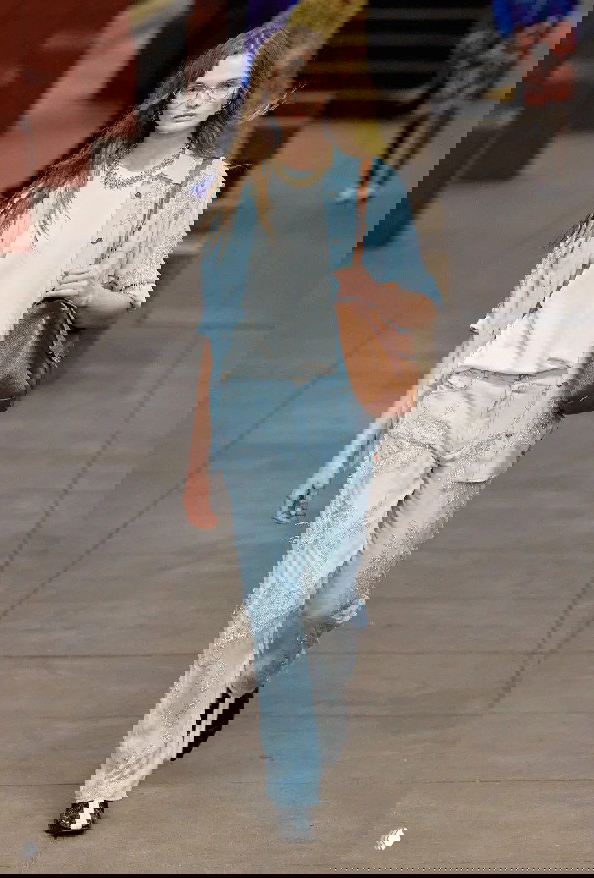 Модель в небесно‑голубом комплекте с вышивкой и сумкой идёт по платформе нью‑йоркского метро в рамках показа Chanel, фон — типичные колонны станции.